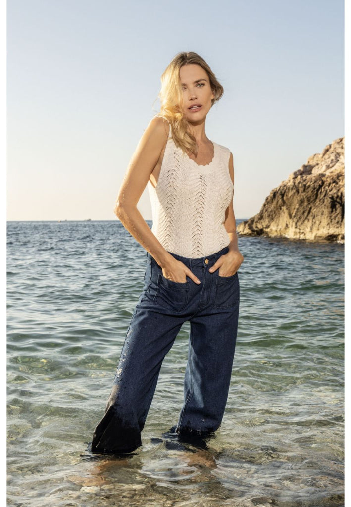 Woman standing in shallow water near a rocky shore wearing a sleeveless top and pants.