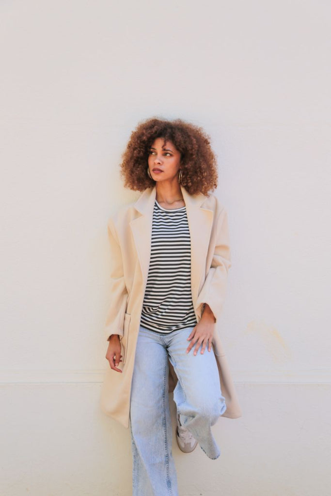 Woman wearing a beige coat, striped shirt, and light blue jeans against a plain background