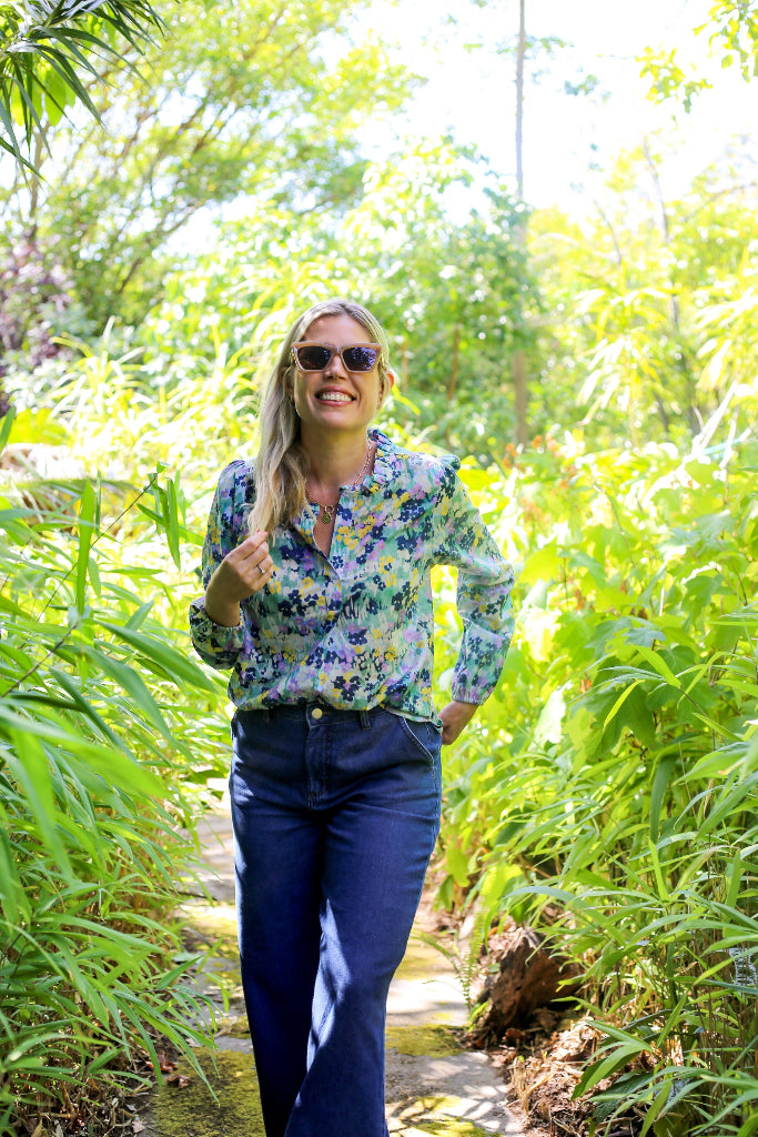 Woman in a floral shirt and sunglasses walking through a lush green forest.