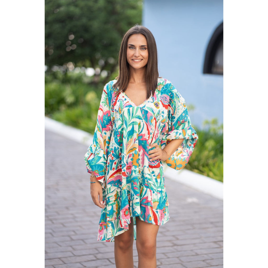 Isla Bonita Dress Woman wearing a colorful floral dress standing on a pathway with greenery and a blue wall in the background.