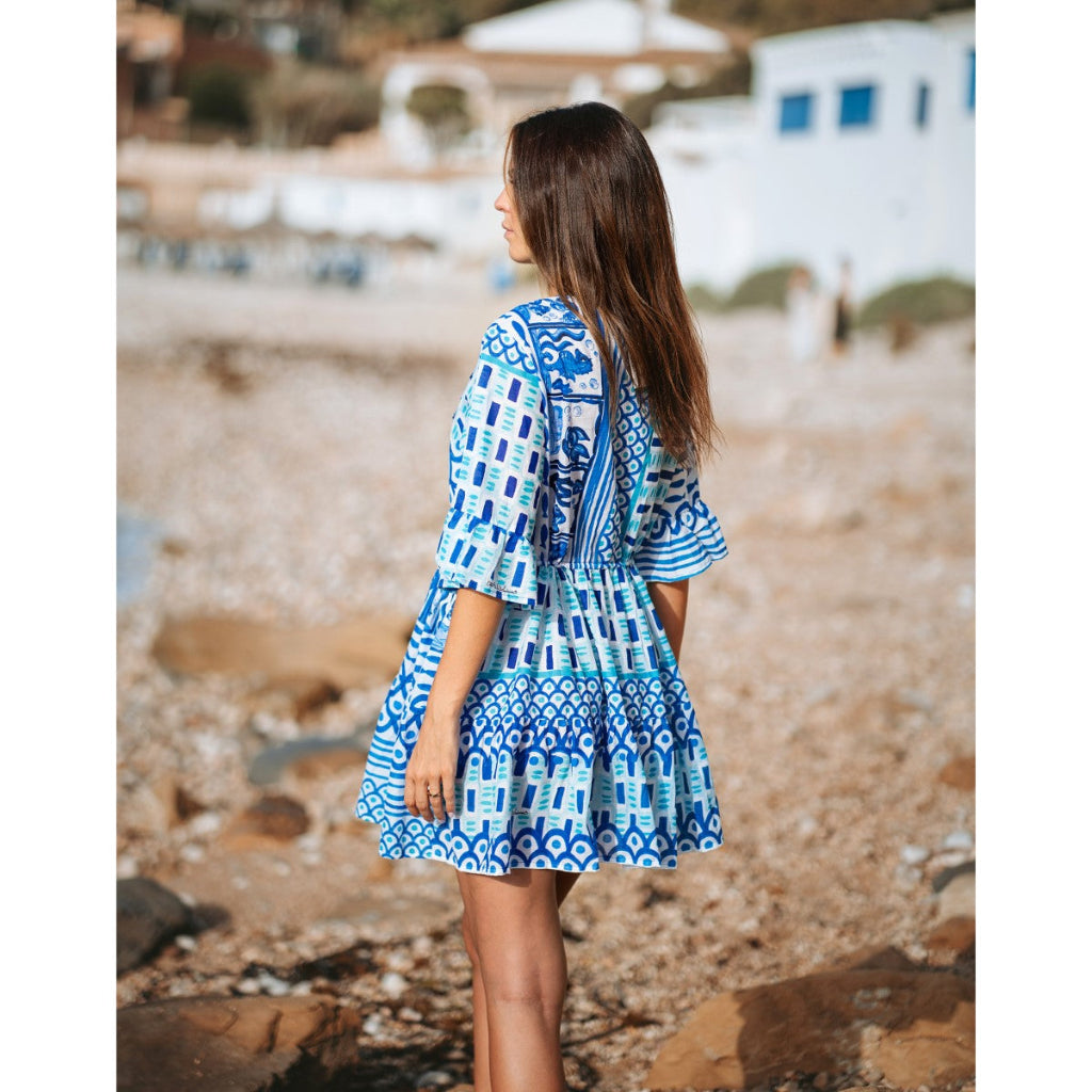 Woman in a blue patterned dress standing on a beach with boats in the background