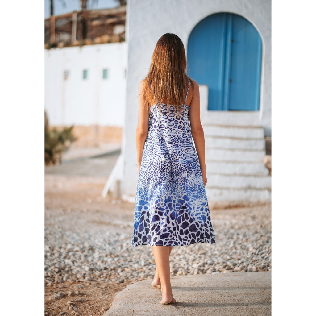 Woman in a blue and white patterned dress walking on a pebbly path with a white building and blue door in the background.