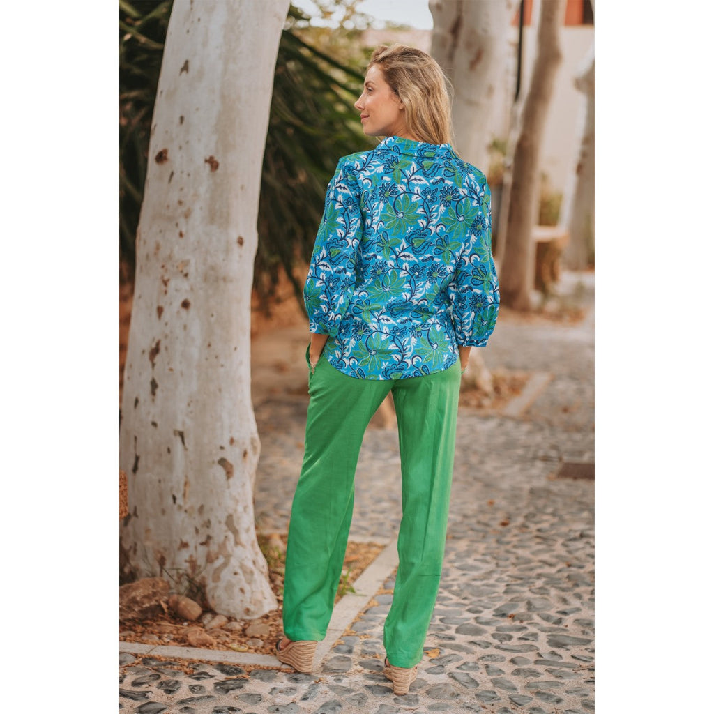 Woman wearing a blue floral blouse and green pants standing on a stone path with trees in the background.