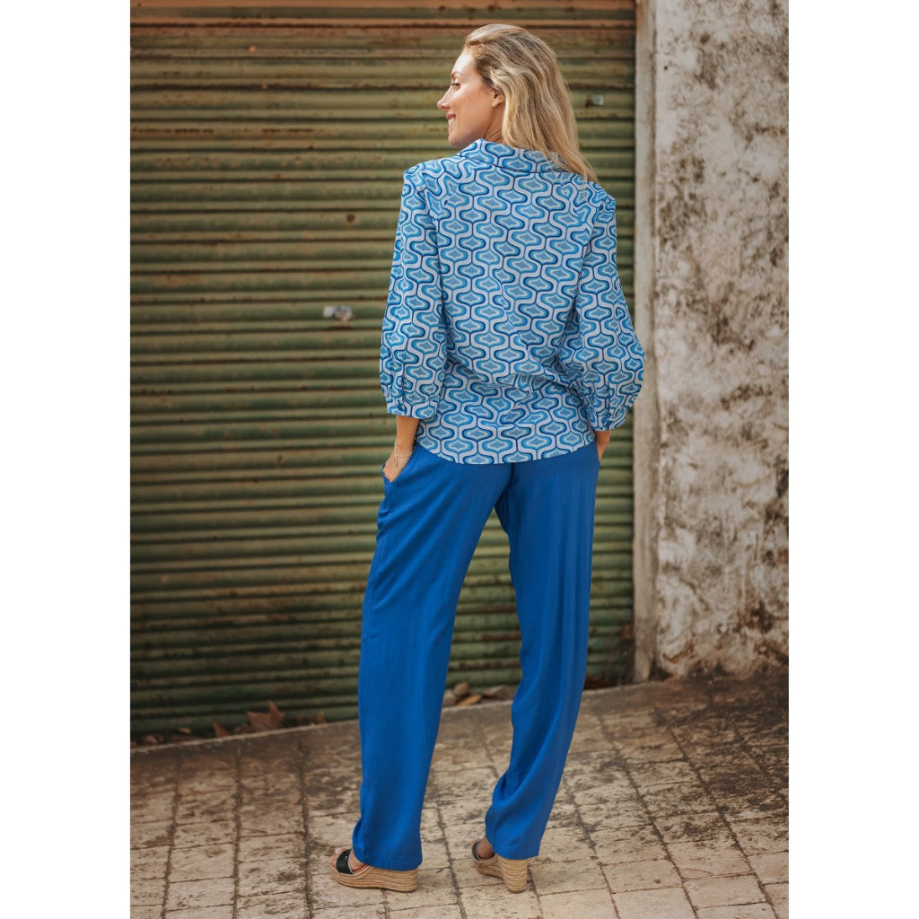 Woman wearing a blue patterned blouse and blue pants standing against a corrugated metal wall.