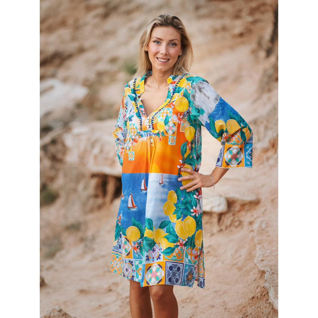 Woman wearing a colorful dress with a pattern of lemons and boats on a rocky beach.
