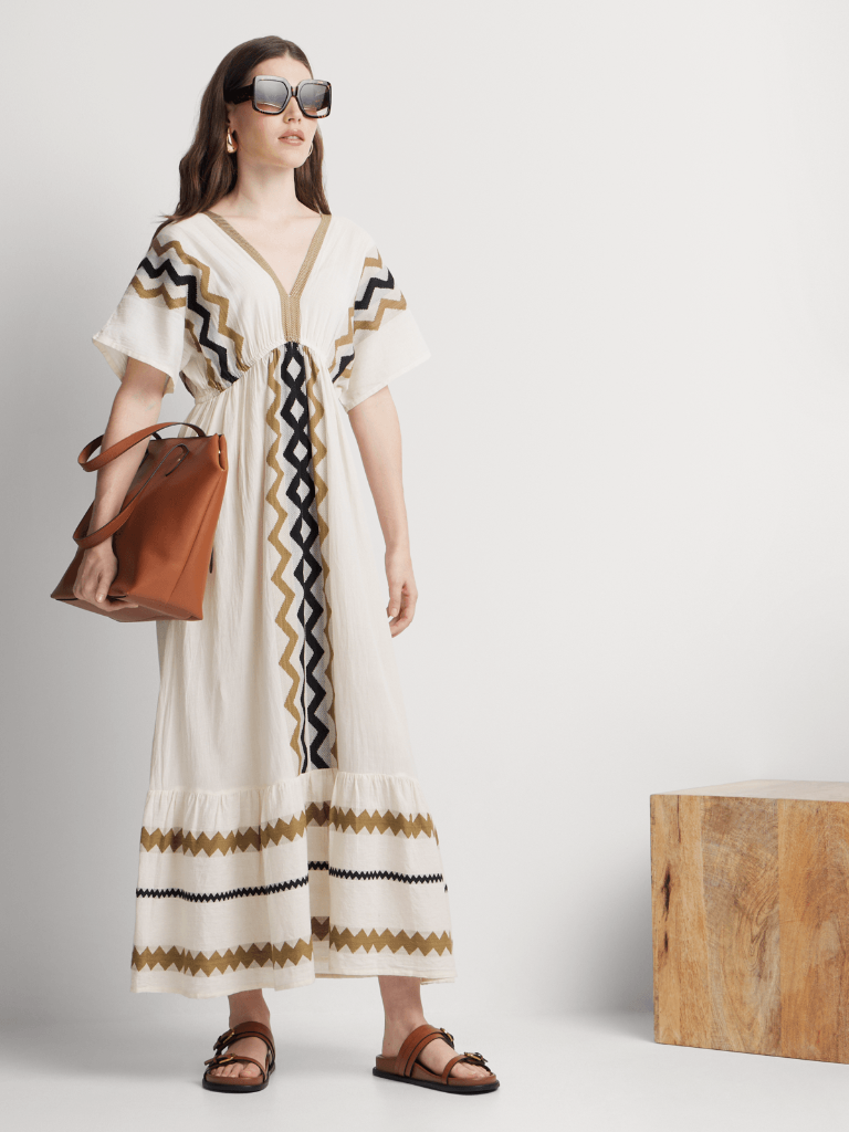 Woman wearing a long, patterned dress with sunglasses and a brown bag on a plain background