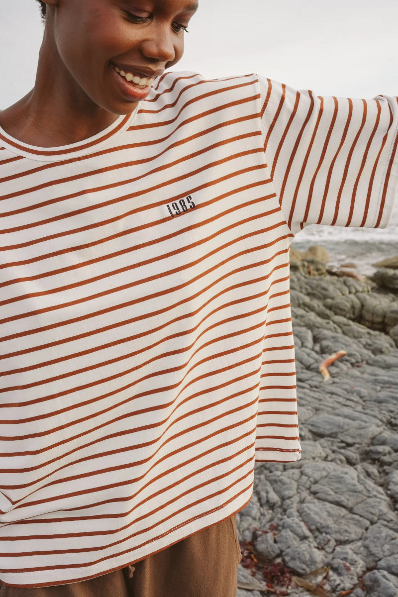 Person wearing a striped shirt with a logo, standing on a rocky beach.