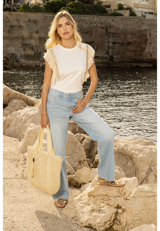 Woman standing on rocks by a body of water wearing a white top and light blue jeans.
