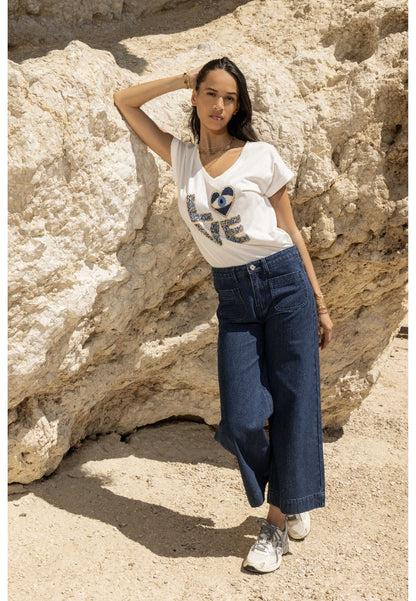 Woman wearing a white t-shirt with blue text and blue jeans standing against a rocky background