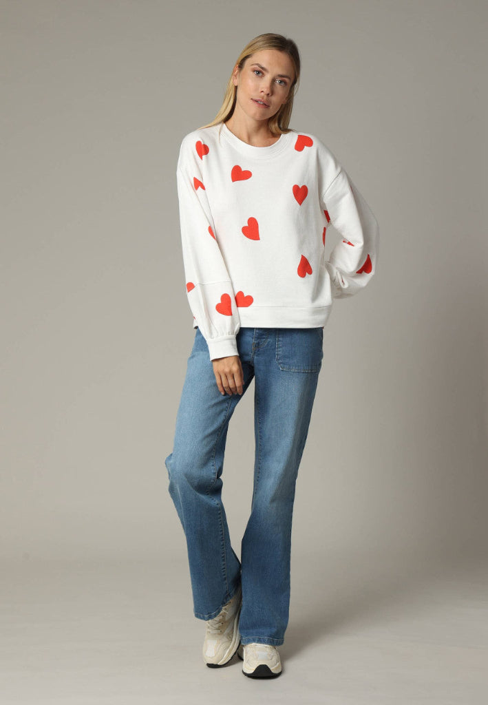 Woman wearing a white sweater with red heart patterns and blue jeans on a plain background