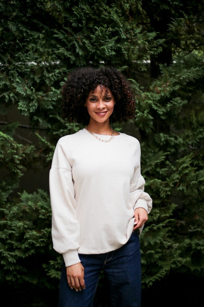 A woman with curly black hair wearing a cream-colored long-sleeve crewneck sweatshirt and dark blue jeans, standing in front of a dense green hedge.