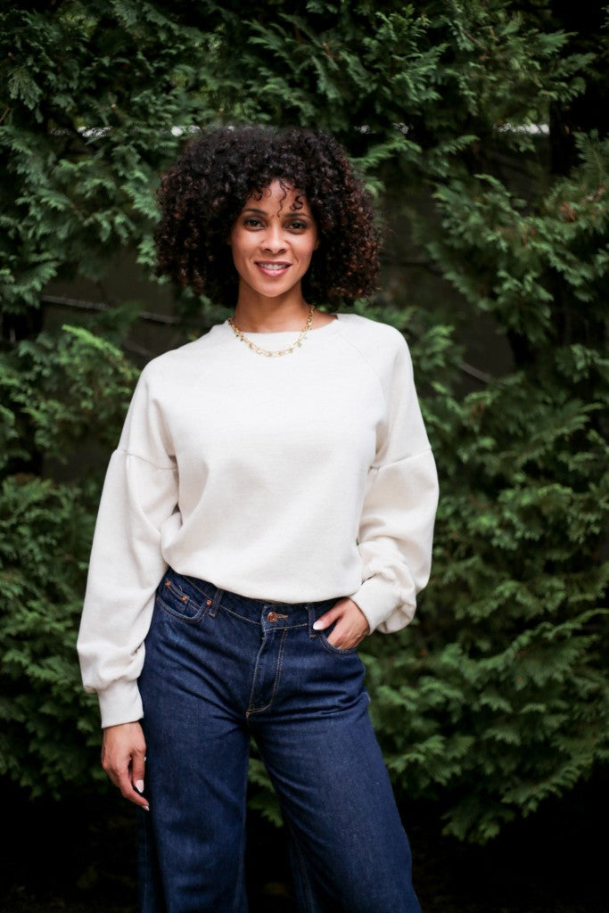 A woman with curly dark hair wearing a cream-colored long-sleeve crewneck sweatshirt and dark blue jeans, standing in front of a background of green foliage.