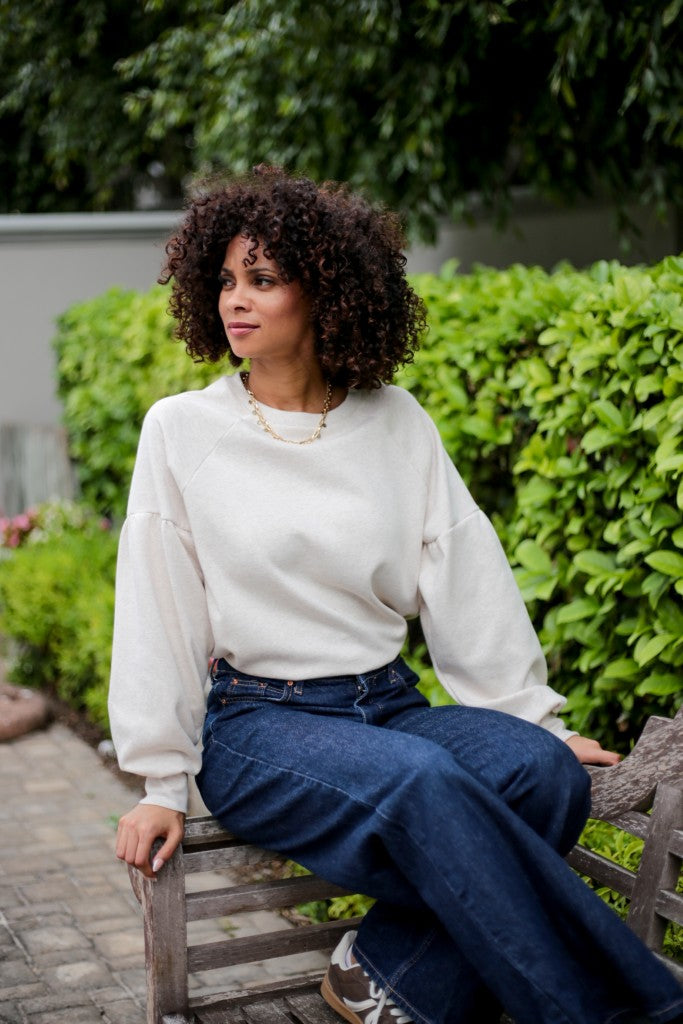 A woman with curly dark hair sits on a wooden bench in a garden setting, wearing a cream-colored long-sleeve crewneck sweatshirt and dark blue wide-leg jeans.
