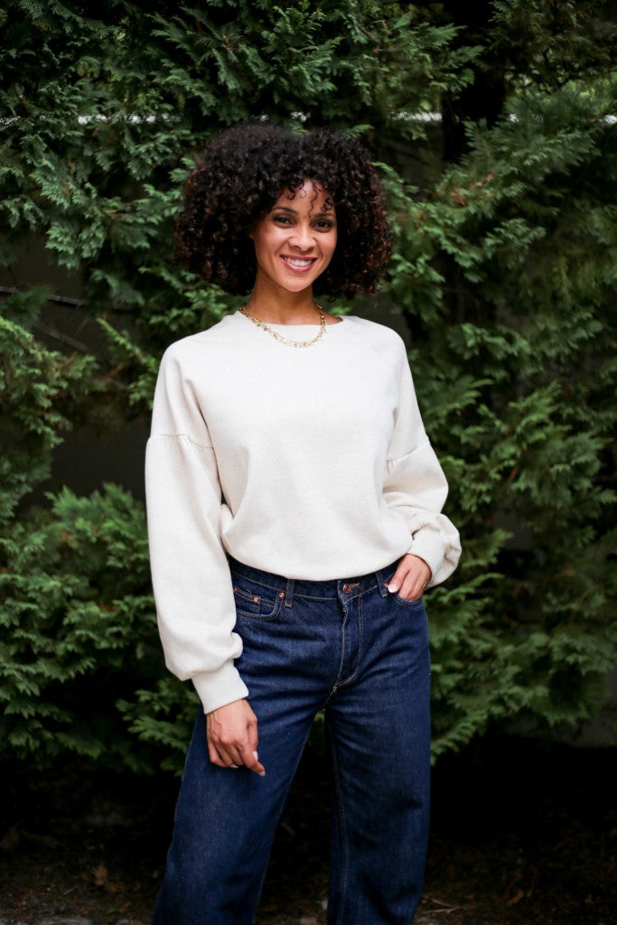 A smiling woman with curly dark hair stands outdoors in front of green trees, wearing a cream-colored long-sleeve crewneck sweatshirt and dark blue jeans.