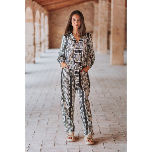 Woman wearing a patterned outfit standing in a sunlit hallway with stone walls.