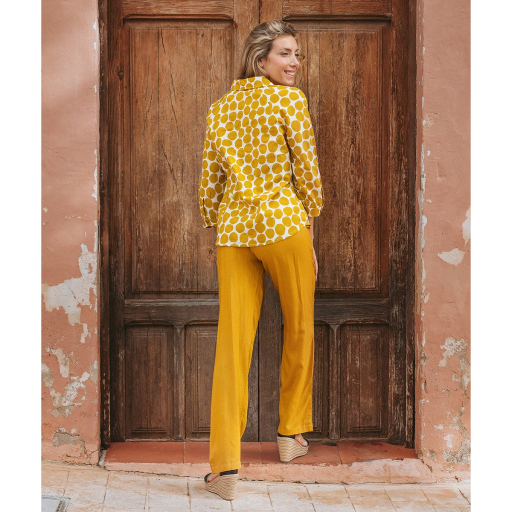 Woman in a yellow polka dot blouse and pants standing in front of a wooden door.