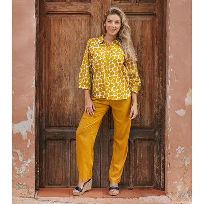Woman wearing a yellow patterned blouse and pants standing in front of a wooden door.