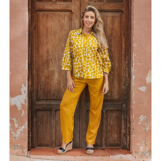 Woman wearing a yellow patterned blouse and pants standing in front of a wooden door.