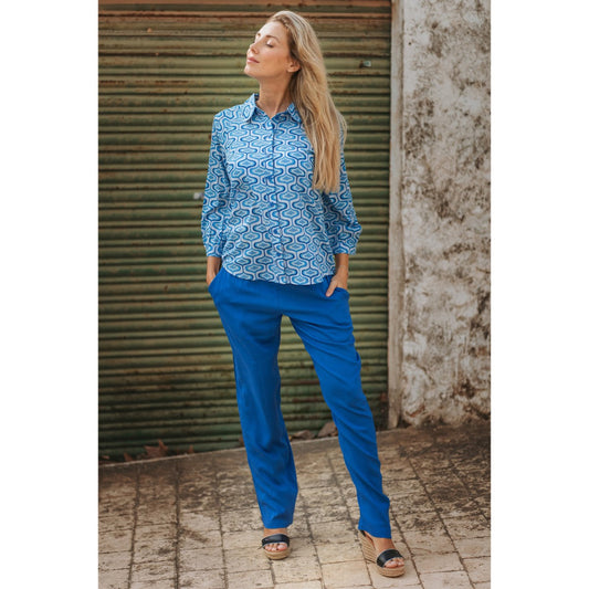 Woman wearing a blue patterned shirt and blue pants standing against a corrugated metal wall.