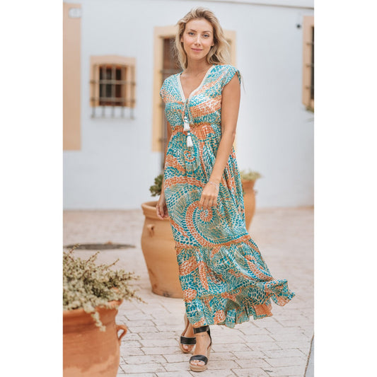 Woman wearing a patterned dress standing outdoors with a white wall and potted plants in the background.