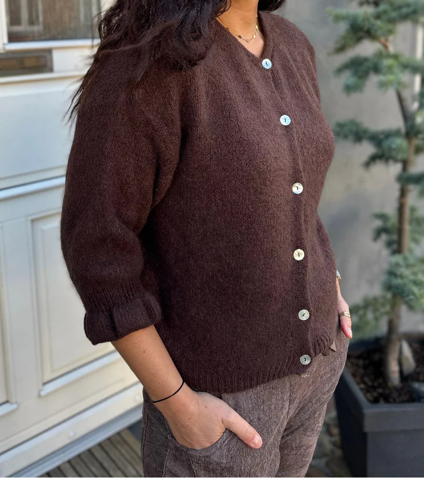 Woman wearing a brown cardigan standing outdoors with a building and plant in the background