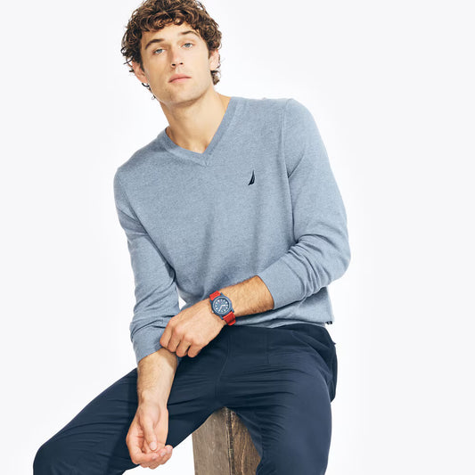 Man wearing a light blue sweater with a small logo, sitting on a wooden stool against a white background.