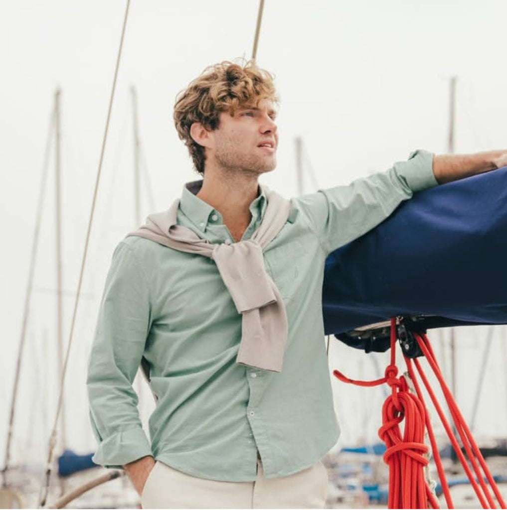 Man on a boat wearing a light green shirt and beige scarf, with a blurred background of water and sky.