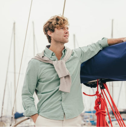 Man on a boat wearing a light green shirt and beige scarf, with a blurred background of water and sky.