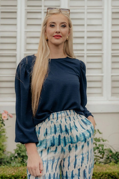 Woman wearing a navy blue top and patterned pants standing in front of a white shutter.