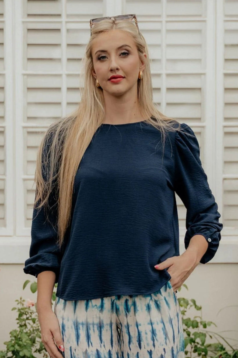 Woman wearing a navy blue top with puffed sleeves in front of a white shuttered window.