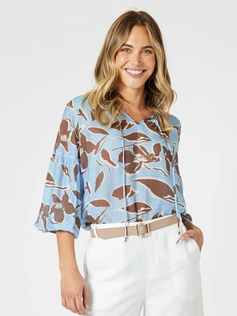 Woman wearing a blue floral blouse with white pants on a plain background
